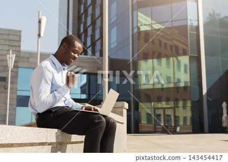 A man sits on a stone bench in front of modern buildings. He uses a laptop and smiles as he works. Sunlight shines on him and the buildings. The scene shows urban life and technology. A man sits on a stone bench in front of modern buildings. He uses a laptop and smiles as he works. Sunlight shines on him and the buildings. The scene shows urban life and technology. 143454417