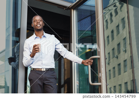 A person holds a coffee cup while standing at the entrance of a modern building. The sun shines on the glass facade, reflecting the surroundings. The location seems busy with activity. A person holds a coffee cup while standing at the entrance of a modern building. The sun shines on the glass facade, reflecting the surroundings. The location seems busy with activity. 143454799