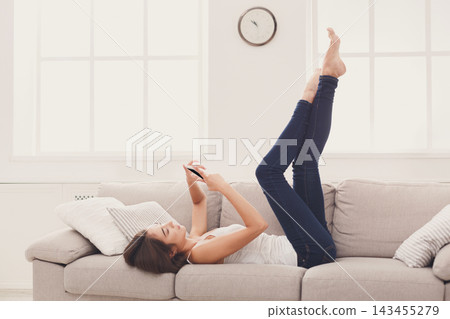 A woman lies on a couch with her legs up while using a smartphone. She is in a well-lit room with a clock on the wall. The atmosphere looks casual and cozy. A woman lies on a couch with her legs up while using a smartphone. She is in a well-lit room with a clock on the wall. The atmosphere looks casual and cozy. 143455279