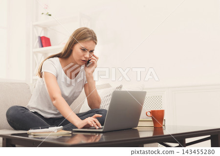 A young woman sits on a couch with a laptop open in front of her. She is holding a phone to her ear while typing. The room is bright with shelves in the background and a cup on the table. A young woman sits on a couch with a laptop open in front of her. She is holding a phone to her ear while typing. The room is bright with shelves in the background and a cup on the table. 143455280