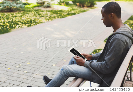 A man sits on a bench in a park, focused on his tablet. He is surrounded by greenery and a path. It is daytime, and he appears engaged with the device in his hands. A man sits on a bench in a park, focused on his tablet. He is surrounded by greenery and a path. It is daytime, and he appears engaged with the device in his hands. 143455430