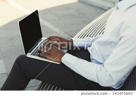 A person types on a laptop while seated on a metal bench. The scene takes place outdoors in a public area with smooth pavement and bright sunlight. A person types on a laptop while seated on a metal bench. The scene takes place outdoors in a public area with smooth pavement and bright sunlight. 143455602
