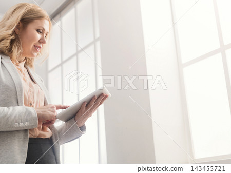 Happy businesswoman at work using digital tablet while standing near window at modern office, copy space Happy businesswoman at work using digital tablet while standing near window at modern office, copy space 143455721