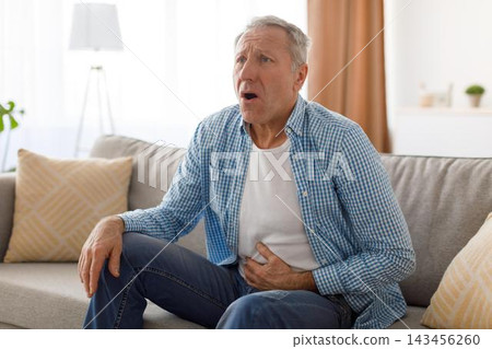 Worried Elderly Caucasian Man Holding His Stomach In Pain While Sitting On Couch In Living Room. Showing Expression Of Discomfort And Concern In Domestic Setting. Healthcare Concept. Worried Elderly Caucasian Man Holding His Stomach In Pain While Sitting On Couch In Living Room. Showing Expression Of Discomfort And Concern In Domestic Setting. Healthcare Concept. 143456260