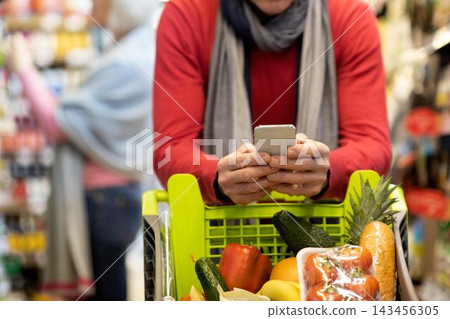 Focused Unrecognizable Person Holding Smartphone In Supermarket Cart Filled With Fresh Groceries, Surrounded By Various Food Products In A Busy Grocery Store Setting. Shopping Concept. Focused Unrecognizable Person Holding Smartphone In Supermarket Cart Filled With Fresh Groceries, Surrounded By Various Food Products In A Busy Grocery Store Setting. Shopping Concept. 143456305