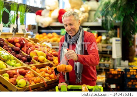 Happy Senior Caucasian Man Enjoying Shopping For Fresh Fruits At Supermarket, Holding Mandarin In One Hand While Selecting Produce. Healthy Lifestyle And Nutrition Focus At Grocery Store. Happy Senior Caucasian Man Enjoying Shopping For Fresh Fruits At Supermarket, Holding Mandarin In One Hand While Selecting Produce. Healthy Lifestyle And Nutrition Focus At Grocery Store. 143456313