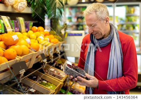 Happy Senior Caucasian Man Examining Packaged Grapes While Shopping For Fresh Fruits At Grocery Store. Enjoying Healthy Eating Choices, Exploring Organic Options For Nutrition In Modern Market. Happy Senior Caucasian Man Examining Packaged Grapes While Shopping For Fresh Fruits At Grocery Store. Enjoying Healthy Eating Choices, Exploring Organic Options For Nutrition In Modern Market. 143456364