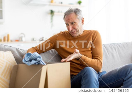 Worried Elderly Caucasian Man Sitting On Couch With Box, Looking Surprised While Cleaning It At Home, Contemplating What's Inside. Concept Of Home Organization And Unboxing Experience. Worried Elderly Caucasian Man Sitting On Couch With Box, Looking Surprised While Cleaning It At Home, Contemplating What's Inside. Concept Of Home Organization And Unboxing Experience. 143456365