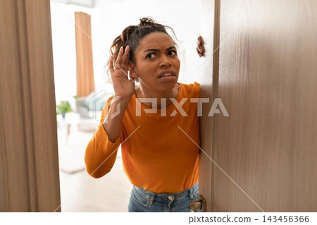 Worried Black Female Listening At Door, Looking Concerned With Hand On Ear, In Home Setting. Emotion Clearly Visible As She Tries To Hear What Is Happening Outside, Indoor Home Environment. Worried Black Female Listening At Door, Looking Concerned With Hand On Ear, In Home Setting. Emotion Clearly Visible As She Tries To Hear What Is Happening Outside, Indoor Home Environment. 143456366