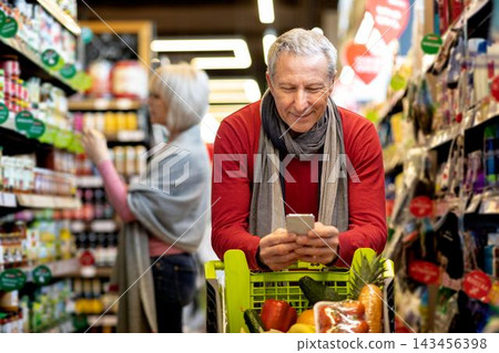 Smiling Elderly Caucasian Man Using Smartphone While Shopping For Fresh Produce In Supermarket, Enjoying His Time At Store. Senior Man Engaged In Grocery Shopping Experience, Weight Loss Concept. Smiling Elderly Caucasian Man Using Smartphone While Shopping For Fresh Produce In Supermarket, Enjoying His Time At Store. Senior Man Engaged In Grocery Shopping Experience, Weight Loss Concept. 143456398