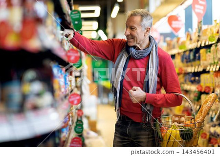 Smiling Elderly Caucasian Man Looking At Fresh Produce While Shopping In Supermarket, Holding A Basket With Groceries. Enjoying His Time Selecting Healthy Foods In Retail Environment. Lifestyle 143456401