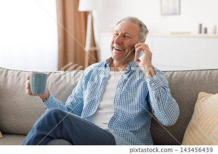 Happy Elderly Caucasian Man Smiling While Conversing On Phone And Holding A Cup Of Coffee, Sitting Comfortably At Home On Sofa. Enjoying Relaxing Moment And Social Connection, Lifestyle Concept. Happy Elderly Caucasian Man Smiling While Conversing On Phone And Holding A Cup Of Coffee, Sitting Comfortably At Home On Sofa. Enjoying Relaxing Moment And Social Connection, Lifestyle Concept. 143456439