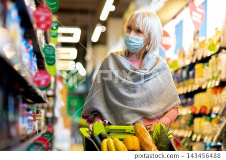 Focused Senior Caucasian Woman Wearing Mask While Shopping For Fresh Fruits And Vegetables In Grocery Store, Standing Next To Shelves Filled With Products. Healthy Lifestyle Concept. Focused Senior Caucasian Woman Wearing Mask While Shopping For Fresh Fruits And Vegetables In Grocery Store, Standing Next To Shelves Filled With Products. Healthy Lifestyle Concept. 143456488