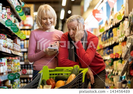 Worried Elderly Caucasian Man With Gray Hair, Resting Head On Hand While Happy Woman, Mid Age, Writing Shopping List In Grocery Store. Supermarket Shopping Concept. Worried Elderly Caucasian Man With Gray Hair, Resting Head On Hand While Happy Woman, Mid Age, Writing Shopping List In Grocery Store. Supermarket Shopping Concept. 143456489