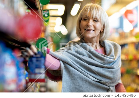 Smiling Elderly Caucasian Woman Shopping In Supermarket. She Is Selecting Items From The Shelf, Enjoying The Shopping Experience In A Bright, Colorful Store Setting. Smiling Elderly Caucasian Woman Shopping In Supermarket. She Is Selecting Items From The Shelf, Enjoying The Shopping Experience In A Bright, Colorful Store Setting. 143456490