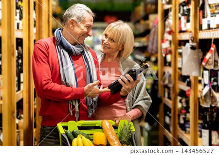 Smiling Elderly Caucasian Man And Woman Enjoying Shopping Together At Supermarket, Holding A Bottle Of Wine In A Cart While Choosing Fresh Produce, Happy Moments In Grocery Store. Smiling Elderly Caucasian Man And Woman Enjoying Shopping Together At Supermarket, Holding A Bottle Of Wine In A Cart While Choosing Fresh Produce, Happy Moments In Grocery Store. 143456527