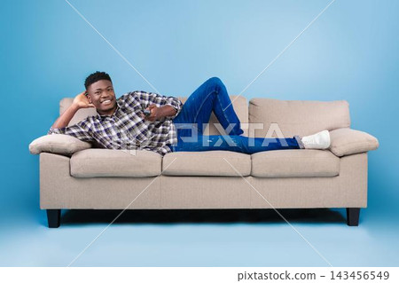 Smiling Black Male Relaxing On Couch At Home, Lying Comfortably With One Arm Behind His Head, Looking At Camera, In Cozy Living Room Setting. Casual Lifestyle And Comfort Concept. Smiling Black Male Relaxing On Couch At Home, Lying Comfortably With One Arm Behind His Head, Looking At Camera, In Cozy Living Room Setting. Casual Lifestyle And Comfort Concept. 143456549