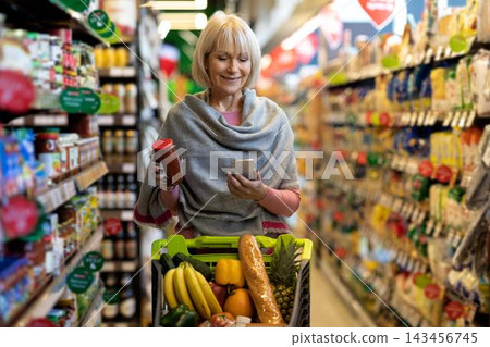 Smiling Elderly Caucasian Woman Holding Smartphone And Jar While Shopping In Supermarket, Picking Fresh Fruits And Vegetables, Enjoying Healthy Lifestyle In Grocery Store Environment. Smiling Elderly Caucasian Woman Holding Smartphone And Jar While Shopping In Supermarket, Picking Fresh Fruits And Vegetables, Enjoying Healthy Lifestyle In Grocery Store Environment. 143456745