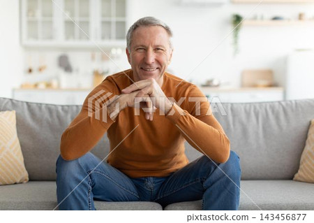 Smiling Mature Caucasian Man Seated Cross-legged On Couch In Cozy Living Room, Exuding Happiness And Contentment, Engaging In Relaxation And Enjoyment At Home. Family Lifestyle Concept. Smiling Mature Caucasian Man Seated Cross-legged On Couch In Cozy Living Room, Exuding Happiness And Contentment, Engaging In Relaxation And Enjoyment At Home. Family Lifestyle Concept. 143456877