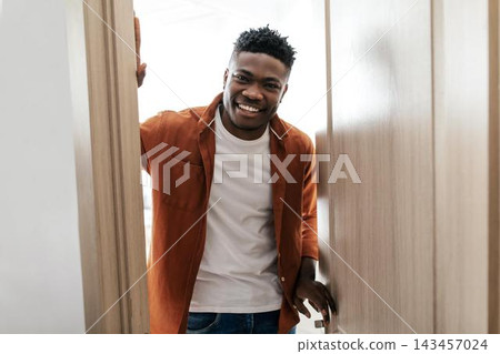 Smiling Young Black Man Standing At Doorway In Bright Modern Home, Engaging Friendly Gesture And Inviting Expression. Indoor Setting With Joyful Ambiance, Home Lifestyle Concept. Smiling Young Black Man Standing At Doorway In Bright Modern Home, Engaging Friendly Gesture And Inviting Expression. Indoor Setting With Joyful Ambiance, Home Lifestyle Concept. 143457024