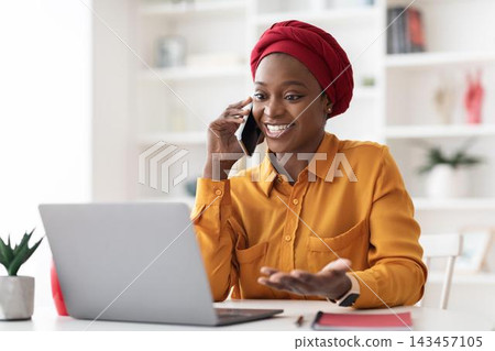 Happy Black Woman Engaged On Phone Call While Using Laptop, Smiling And Communicating From Home Office, Demonstrating Professional Success And Modern Business Concept. 143457105