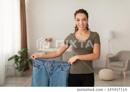 Smiling Young Woman Holding Oversized Jeans, Celebrating Weight Loss Achievement At Home In Modern Living Area. Positive Emotion Reflected As She Shows Progress In Her Fitness Journey. Weight Loss Smiling Young Woman Holding Oversized Jeans, Celebrating Weight Loss Achievement At Home In Modern Living Area. Positive Emotion Reflected As She Shows Progress In Her Fitness Journey. Weight Loss 143457114