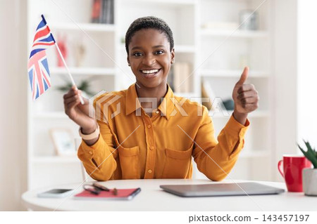 Smiling Black Woman Showing Joy While Holding British Flag In Modern Office Space, Giving Thumbs Up. Expressing Happiness And Success In Professional Environment, Business Concept. 143457197