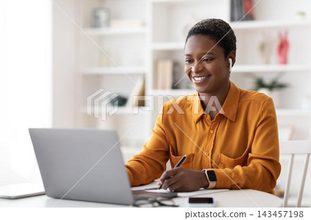 Smiling Black Woman Focused While Working On Laptop At Home Office, Taking Notes And Engaging With Digital Content. Young Adult Female Enjoying Productive Day In Comfortable Workspace. Smiling Black Woman Focused While Working On Laptop At Home Office, Taking Notes And Engaging With Digital Content. Young Adult Female Enjoying Productive Day In Comfortable Workspace. 143457198