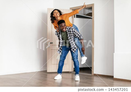 Joyful Couple Enjoying Playful Moment Together In Living Room, Happy Black Man Carrying Smiling Black Woman On His Back While Laughing, Indoor Setting At Home, Fun Lifestyle Concept. Joyful Couple Enjoying Playful Moment Together In Living Room, Happy Black Man Carrying Smiling Black Woman On His Back While Laughing, Indoor Setting At Home, Fun Lifestyle Concept. 143457201