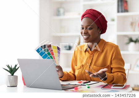 Smiling Black Female Designer Engaged In Creative Work With Color Swatches While Using Laptop At Modern Office Workspace. Happy Professional Focused On Design Project. Business Concept. 143457226
