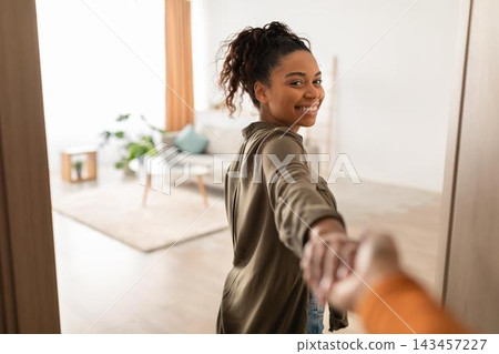 Smiling Black Woman With Curly Hair Joyfully Holding Hand And Turning Around In Bright Modern Living Room, Exuding Happiness And Warmth, Home Lifestyle Concept. Smiling Black Woman With Curly Hair Joyfully Holding Hand And Turning Around In Bright Modern Living Room, Exuding Happiness And Warmth, Home Lifestyle Concept. 143457227