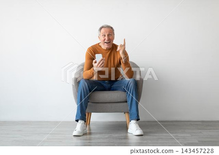 Happy Elderly Caucasian Man Smiling While Using Smartphone And Pointing Up In Living Room At Home. Enjoying Technology And Communication, Excited About Something On Screen. Digital Lifestyle Concept. Happy Elderly Caucasian Man Smiling While Using Smartphone And Pointing Up In Living Room At Home. Enjoying Technology And Communication, Excited About Something On Screen. Digital Lifestyle Concept. 143457228