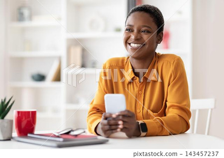 Happy Smiling Black Woman In Office Environment Holding Smartphone And Looking Engaged With Positive Emotion. She Is Enjoying Interaction In Modern Workspace With Clean Design. Happy Smiling Black Woman In Office Environment Holding Smartphone And Looking Engaged With Positive Emotion. She Is Enjoying Interaction In Modern Workspace With Clean Design. 143457237