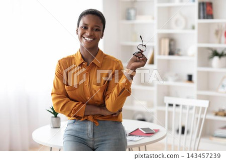 Smiling Black Woman With Short Hair Holding Glasses While Standing In Office Setting, Showing Confidence And Professionalism In Work Environment. Business Concept. Smiling Black Woman With Short Hair Holding Glasses While Standing In Office Setting, Showing Confidence And Professionalism In Work Environment. Business Concept. 143457239