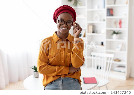 Smiling Black Woman Wearing Glasses And Red Headscarf, Confidently Posing At Home Office, Engaging In Professional Environment, Embracing Empowerment In Her Career. Business Concept. 143457240