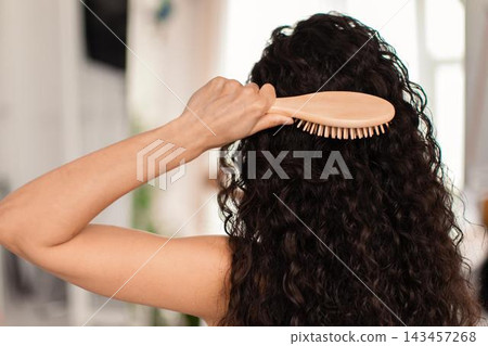 Focused Unrecognizable Woman Brushing Curly Hair With Wooden Brush In Modern Home Interior, Sitting In Cozy Atmosphere Surrounded By Light, Relaxed Space. Hair Care Concept. Focused Unrecognizable Woman Brushing Curly Hair With Wooden Brush In Modern Home Interior, Sitting In Cozy Atmosphere Surrounded By Light, Relaxed Space. Hair Care Concept. 143457268