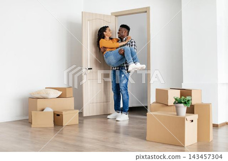 Joyful Emotion. Happy Black Couple In Their 30s Unpacking Boxes While Moving Into New Apartment. Man Lifting Woman With Joyful Expression In Modern Living Room. Moving And Home Concept. Joyful Emotion. Happy Black Couple In Their 30s Unpacking Boxes While Moving Into New Apartment. Man Lifting Woman With Joyful Expression In Modern Living Room. Moving And Home Concept. 143457304