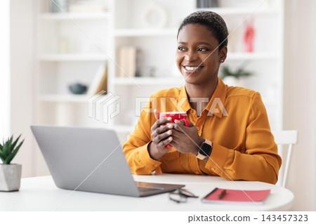 Smiling Black Woman In Yellow Blouse Holding Coffee Mug While Working At Home Office, Sitting At Desk With Laptop, Reflecting Positive Emotion And Professionalism, Business Concept. Smiling Black Woman In Yellow Blouse Holding Coffee Mug While Working At Home Office, Sitting At Desk With Laptop, Reflecting Positive Emotion And Professionalism, Business Concept. 143457323