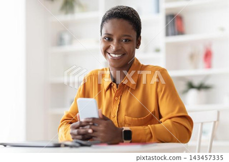 Smiling Black Woman In Her 30s Holding Smartphone While Sitting At A Desk In A Bright Office Environment, Engaged In Work Or Communication. Business Concept. Smiling Black Woman In Her 30s Holding Smartphone While Sitting At A Desk In A Bright Office Environment, Engaged In Work Or Communication. Business Concept. 143457335