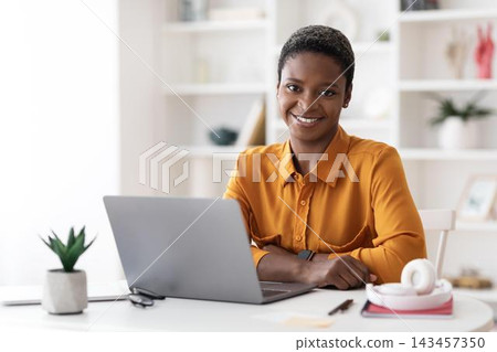 Smiling Black Woman In Her 30s Sitting At Desk With Laptop, Working In Cozy Home Office, Showing Focused Expression While Engaging With Her Work. Business Concept. Smiling Black Woman In Her 30s Sitting At Desk With Laptop, Working In Cozy Home Office, Showing Focused Expression While Engaging With Her Work. Business Concept. 143457350