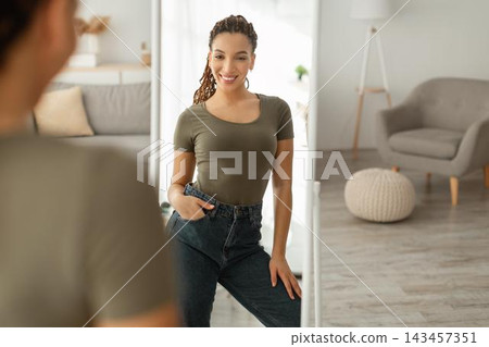 Smiling Young Black Woman Posing Confidently In Front Of Mirror At Home, Wearing Casual Attire. Embracing Self-confidence In Stylish Environment. Fashion Concept. Smiling Young Black Woman Posing Confidently In Front Of Mirror At Home, Wearing Casual Attire. Embracing Self-confidence In Stylish Environment. Fashion Concept. 143457351