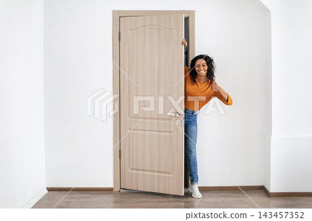 Smiling Black Woman In Casual Outfit Giving Thumbs Up While Peeking Through Open Door In Bright Home Interior. Positive Emotion Displayed In A Welcoming Environment Reflecting Lifestyle Concept. Smiling Black Woman In Casual Outfit Giving Thumbs Up While Peeking Through Open Door In Bright Home Interior. Positive Emotion Displayed In A Welcoming Environment Reflecting Lifestyle Concept. 143457352