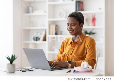 Smiling Black Woman In Her 30s Working At Laptop In Modern Home Office Setting, Focused On Task. Displays Productive Home Environment And Professional Conduct. Business Concept. Smiling Black Woman In Her 30s Working At Laptop In Modern Home Office Setting, Focused On Task. Displays Productive Home Environment And Professional Conduct. Business Concept. 143457353
