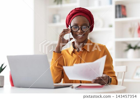 Smiling African American Woman Engaged In Phone Conversation While Reviewing Documents At Her Office Desk, Creating A Productive Work Environment, Embracing Business Concept. Smiling African American Woman Engaged In Phone Conversation While Reviewing Documents At Her Office Desk, Creating A Productive Work Environment, Embracing Business Concept. 143457354