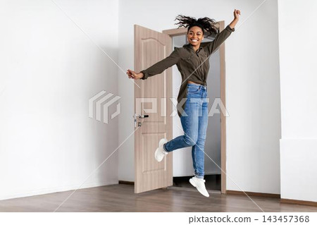 Happy Smiling African American Woman Jumping With Joy In Bright Modern Room At Home, Enjoying Free Space And Positive Vibes. Concept Of Wellness And Happiness In Lifestyle Setting. 143457368