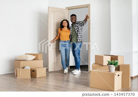 Happy African American Couple Smiling And Unpacking Boxes Together In Their New Home, Standing At Doorway And Enjoying Their New Space, Embracing A Fresh Start And New Beginnings. 143457370