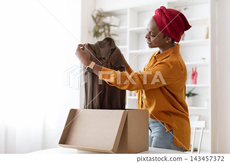 Excited Smiling African American Woman Unboxing New Clothes With Joy In Home Office Setting, Holding A Brown Shirt While Standing By The Table. Unboxing And Shopping Concept. 143457372