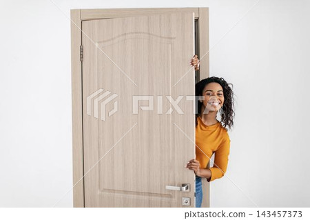 Smiling Young Black Woman Joyfully Peeking Through An Open Door At Home, Creating A Welcoming Atmosphere In A Cozy Interior Setting. Cheerful Emotion Reflected In Her Face. 143457373