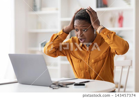 Worried Black Woman In Her 30s Sitting At Desk In Modern Office, Holding Hands On Head While Staring At Laptop, Expressing Stress Over Work. Business Concept. 143457374