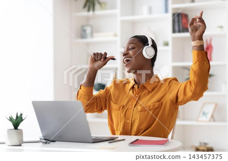 Smiling Black Female Freelancer Enjoying Music While Working On Laptop At Home Office, Engaging In Creative Tasks. Cheerful Atmosphere In Stylish, Modern Workspace, Freelance Lifestyle. Smiling Black Female Freelancer Enjoying Music While Working On Laptop At Home Office, Engaging In Creative Tasks. Cheerful Atmosphere In Stylish, Modern Workspace, Freelance Lifestyle. 143457375